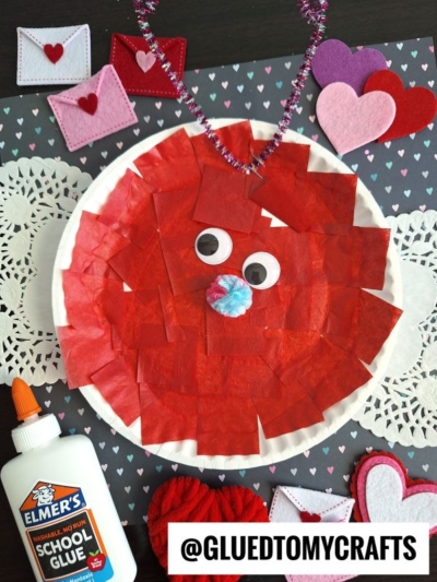 A red tissue paper paper plate lovebug with googly eyes, blue pom-pom nose, and pipe cleaner antennae sits on a paper plate, surrounded by heart decorations, glue, and doilies.