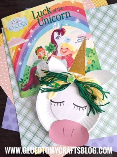 A children’s book titled “Luck of the Unicorn” sits beside a paper plate unicorn craft, complete with yarn hair, closed eyes, a gold horn, and paper ears, all set atop colorful patterned papers.