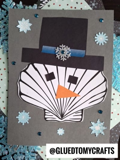 A craft card featuring a seashell snowman made from a paper seashell as the face, with a black paper hat, orange triangle nose, and square eyes. Blue snowflakes and gems decorate the gray background. Text: @GLUEDTOMYCRAFTS.