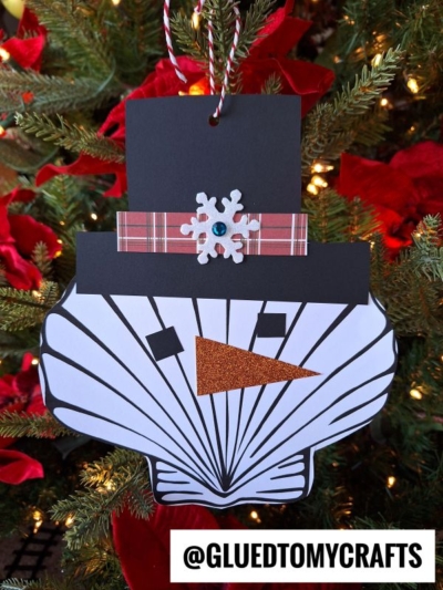 A handmade seashell snowman ornament made from a painted scallop shell, black paper hat, plaid ribbon with a snowflake, and an orange glitter paper nose, hanging on a Christmas tree.