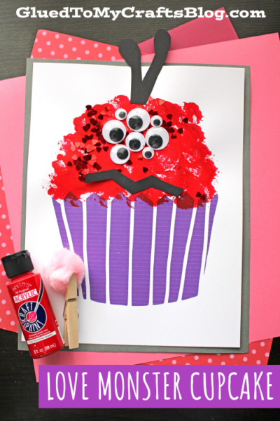 A handmade “Love Monster Cupcake” craft features a red, textured cupcake top inspired by a pom pom love monster, decorated with googly eyes and heart-shaped confetti, purple cupcake liner, and black paper antennae. Supplies and paint sit beside the artwork.