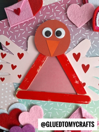 A handmade popsicle stick love bird Valentine’s craft made from red paper, googly eyes, an orange beak, and a red popsicle stick triangle, surrounded by felt and paper hearts and small red heart cutouts on a patterned background.