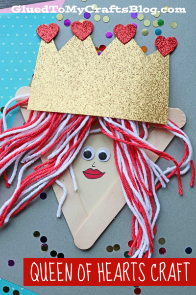 A popsicle stick queen craft shaped like a queen’s face, with red and white yarn hair, a gold glitter crown adorned with red hearts, googly eyes, and the text QUEEN OF HEARTS CRAFT.