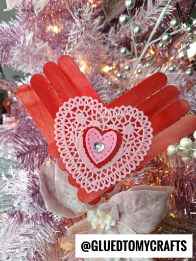 A red heart-shaped ornament made from painted craft sticks and decorated with a pink doily and heart, perfect for learning how to make a heart out of popsicle sticks. Hanging on a white and pink Christmas tree. Label reads @GLUEDTOMYCRAFTS.