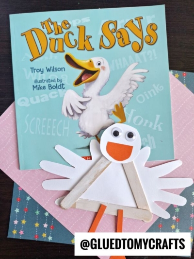 A children’s book titled The Duck Says is displayed with a duck illustration on the cover. In front, a popsicle stick duckling craft made from popsicle sticks, paper, and googly eyes sits on colorful patterned papers.