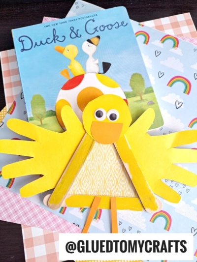 A children’s book titled Duck & Goose is placed behind a popsicle stick duckling craft made from paper, popsicle sticks, and googly eyes. The background features colorful patterned papers with rainbows and hearts.