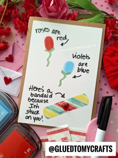 A handmade thumbprint valentine card features two flowers labeled “roses are red” and “violets are blue.” Below, a colorful paper bandage and the text “Here’s a bandaid because I’m stuck on you!” with craft supplies all around.