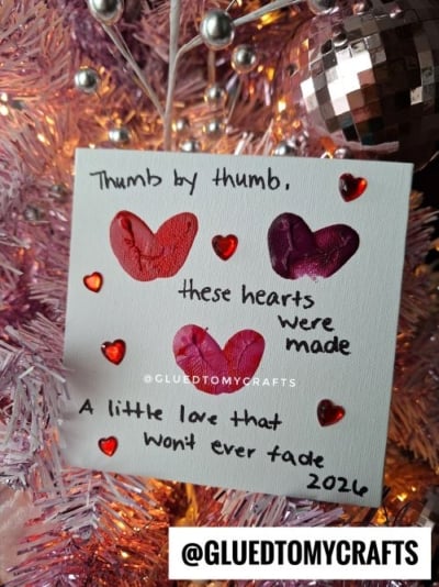 A small canvas with painted thumbprint hearts and red gem stickers hangs on a pink Christmas tree. Like a fingerprint valentine card, it says, Thumb by thumb, these hearts were made. A little love that won’t ever fade. 2026. @GLUEDTOMYCRAFTS.
