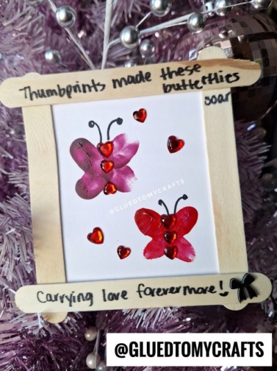 A handmade frame with a butterfly art piece made from thumbprints and heart stickers, just like a creative valentine card kid craft. The frame says, Thumbprints made these buttetties soar, carrying love forevermore! Displayed on a pink Christmas tree.