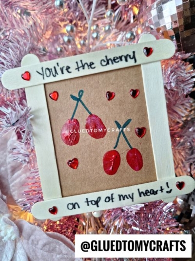 A handmade cherry-themed valentine card kid craft, framed with wooden sticks and red heart stickers. The text reads “YOU’RE the cherry on top of my heart!” on a pink festive background. Instagram: @GLUEDTOMYCRAFTS.