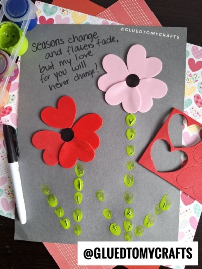 A charming thumbprint valentine card features two paper flowers—one pink, one red—on gray paper. Green paint forms stems and leaves. Nearby craft supplies surround the heartfelt message: “Seasons change and flowers fade, but my love for you will never change!”.