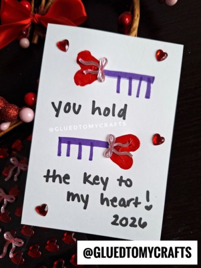A handmade thumbprint card for Valentine's Day features two keys crafted from a purple comb and red heart, with a note reading "You hold the key to my heart! 2026." Red heart gems decorate the corners for an extra special touch.