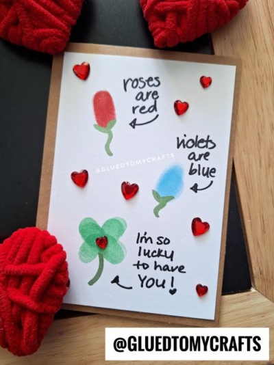 A handmade fingerprint valentine card with thumbprint art showing a red rose and blue heart, surrounded by red gem hearts. The card reads: roses are red, violets are blue, and I'm so lucky to have you!.