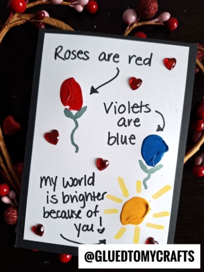 A handmade thumbprint card with painted red and blue flowers, a yellow sun, and heart gems. Text reads: Roses are red, Violets are blue, my world is brighter because of you! @GLUEDTOMYCRAFTS.