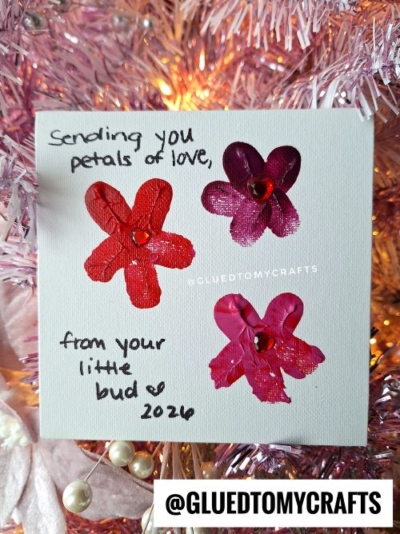 A handmade thumbprint card features three flower shapes crafted from painted red and purple fingerprints with a gem center. This fingerprint valentine card reads: Sending you petals of love, from your little bud ♥ 2024.