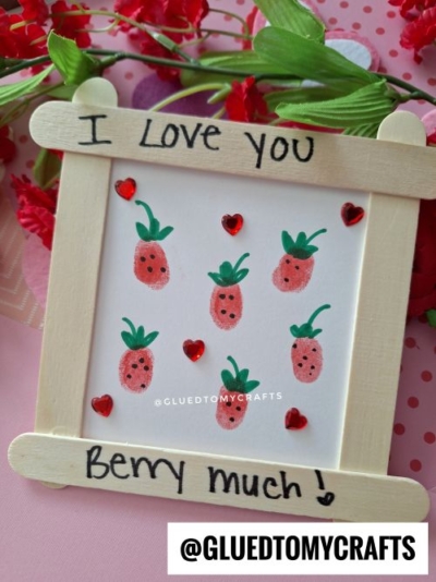 A handmade craft frame with I Love You Berry much! written on it, displaying strawberry finger-paint art and red heart stickers—perfect as a thumbprint valentine card. Red artificial flowers brighten the background.