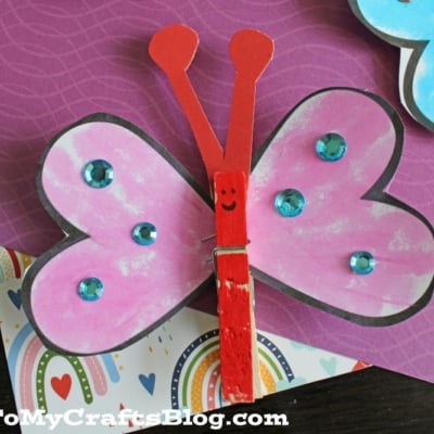 A colorful paper butterfly craft with pink wings, blue gem decorations, and a red love bug valentine clothespin body with a smiling face, displayed on patterned paper backgrounds.