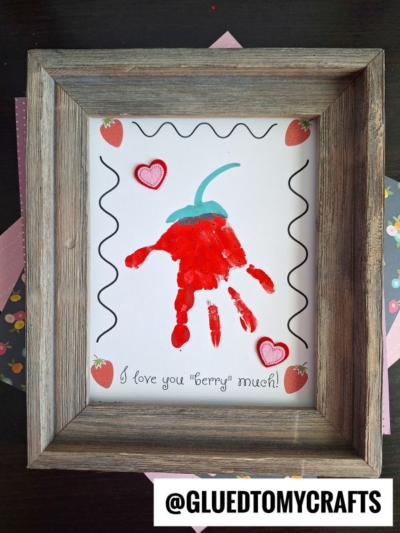 Framed artwork features a red handprint painted like a strawberry with hearts, strawberries, and the sweet phrase "love you berry much!" on white paper. @GLUEDTOMYCRAFTS is at the bottom.