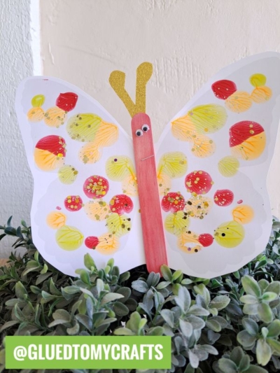 A colorful paint smash butterfly craft made from a painted paper cutout with red, yellow, and orange dots, a pink craft stick body, googly eyes, gold antennae, and displayed on green leaves. @GLUEDTOMYCRAFTS is written at the bottom.