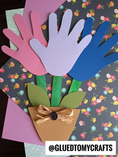 Three colorful paper handprints (pink, purple, blue) form a flower bouquet handprint with green stems and leaves in a brown paper pot, decorated with a bow and button, on patterned background paper—a perfect Mother's Day craft.