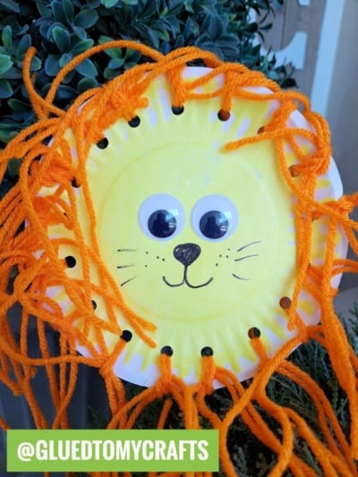 A paper plate yarn lion craft featuring googly eyes, a drawn nose and whiskers, and an orange yarn mane sits among green plants. @GLUEDTOMYCRAFTS appears in a green box at the bottom left.