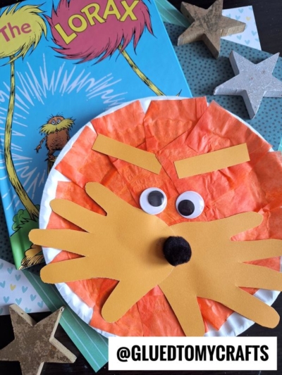 A paper plate Lorax craft, featuring orange tissue paper, paper whiskers, googly eyes, and a pom-pom nose, rests atop a copy of The Lorax book, surrounded by decorative stars.