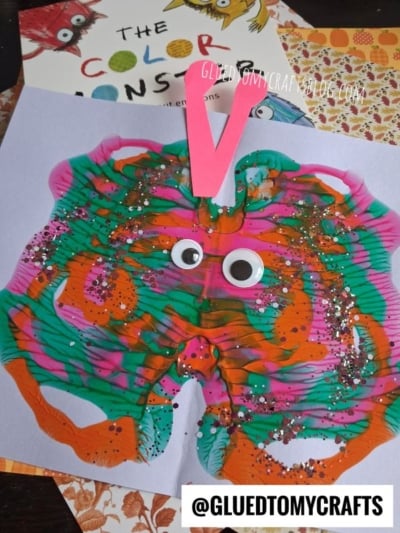 Colorful paint butterfly craft with googly eyes, pink paper antennae, and glitter on a light purple sheet—reminiscent of playful puffy paint splat monsters. A book titled The Color Monster is partly visible in the background.