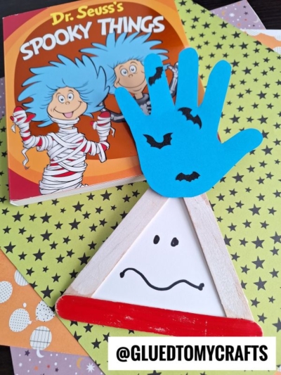 A Dr. Seuss’s “Spooky Things” book lies on patterned papers next to a ghost craft and playful Thing 1 and 2 popsicle sticks with bat cutouts, on top of a white triangle with a sad face and red strip. @GLUEDTOMYCRAFTS is at the bottom.