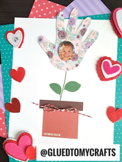 A flower pot handprint craft features a hand-shaped paper cutout with a child's photo in the palm, attached above a paper pot. Drawn stem and leaves connect them, with felt and paper hearts in the background. @GLUEDTOMYCRAFTS is at the bottom.
