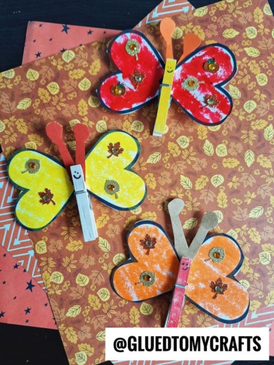 Three colorful butterfly crafts, inspired by a love bug clothespin craft, are made from painted clothespins and paper wings. The red, yellow, and orange butterflies sparkle with glittery decorations and drawn-on smiles. @GLUETOMYCRAFTS is at the bottom.