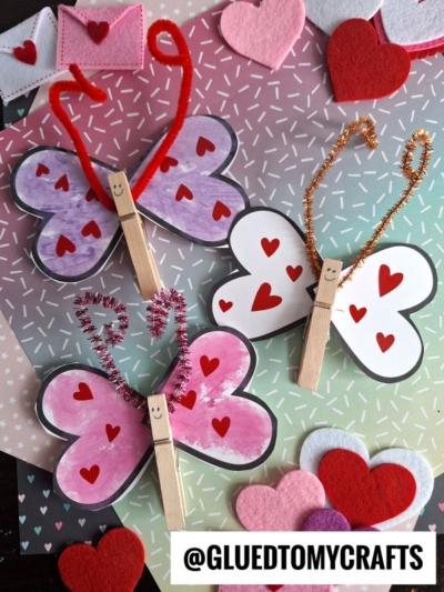 Handmade butterfly crafts made with love bug valentine clothespins, featuring paper wings adorned with red hearts, colorful pipe cleaner antennae, and smiling faces. Displayed on a pastel background with felt hearts for extra charm.