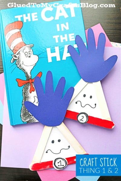 A copy of “The Cat in the Hat” lies on colored paper with two Popsicle Stick Thing 1 & 2 crafts, featuring paper hands and drawn faces labeled as Thing 1 and 2 for a playful book-inspired display.