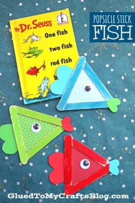 A Dr. Seuss book titled One Fish, Two Fish, Red Fish, Blue Fish sits on a blue background, surrounded by popsicle stick one fish crafts with googly eyes and paper fins.