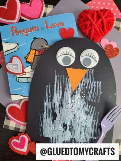 A children’s book titled “Penguin in Love,” heart-shaped decorations, and a fork painted penguin kid craft with googly eyes and an orange beak. Its belly is white, and a purple fork sits beside this adorable penguin creation.