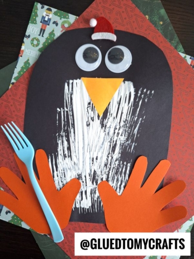 A festive paper penguin craft with orange handprint feet and wings, googly eyes, a Santa hat, yellow beak, and a white belly painted with a blue fork makes the perfect penguin kid craft on red and green holiday paper.
