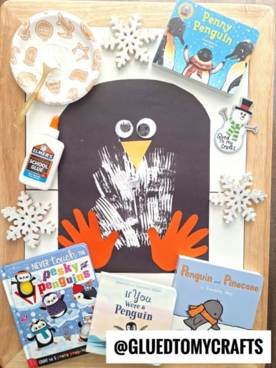 A handmade paper penguin craft featuring a black oval, white painted handprint, orange paper beak and feet, surrounded by penguin books, snowflakes, googly eyes, glue, and a Glued to My Crafts label—a perfect penguin kid craft!.