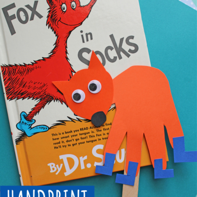 A handmade handprint fox in socks craft with googly eyes sits atop the Fox in Socks Dr. Seuss book, layered on colorful paper. A Handprint Fox in Socks label decorates the corner—perfect for fun fox in socks crafts!.
