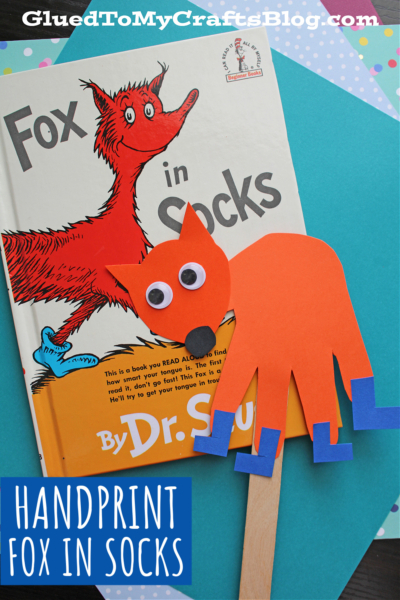 A handmade handprint fox in socks craft with googly eyes sits atop the Fox in Socks Dr. Seuss book, layered on colorful paper. A Handprint Fox in Socks label decorates the corner—perfect for fun fox in socks crafts!.