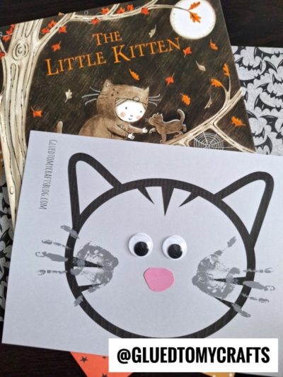 A children's book titled The Little Kitten lies behind a handmade card featuring handprint cat whiskers, googly eyes, and a pink nose. The card rests on patterned paper with a @GLUEDTOMYCRAFTS label.