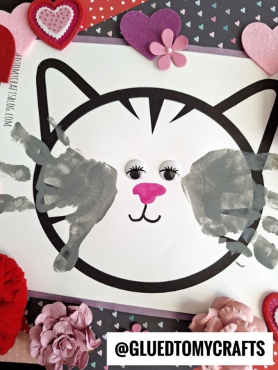 A cute cat face drawing with handprint cat whiskers, gray handprints for cheeks, googly eyes, and a pink nose, surrounded by heart and flower decorations on a colorful background. @GLUEDTOMYCRAFTS is labeled at the bottom.