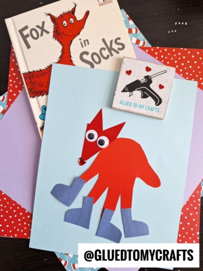 A children’s book “Fox in Socks” with a red handprint fox in socks paper craft wearing blue socks on light blue paper, surrounded by colorful patterned paper. A wooden sign with a glue gun and “Glued To My Crafts” logo is also visible.