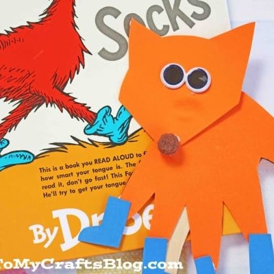 A handmade handprint fox in socks puppet with googly eyes and blue boots sits beside the book Fox in Socks by Dr. Seuss. This adorable fox in socks crafts project is mounted on a wooden stick for storytelling fun.