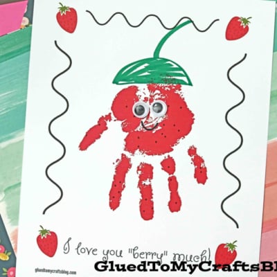 A craft featuring a red handprint decorated as a strawberry with googly eyes, green leaf top, and small strawberry drawings. Text below reads, handprint you love berry much! with colorful paper and confetti in the background.