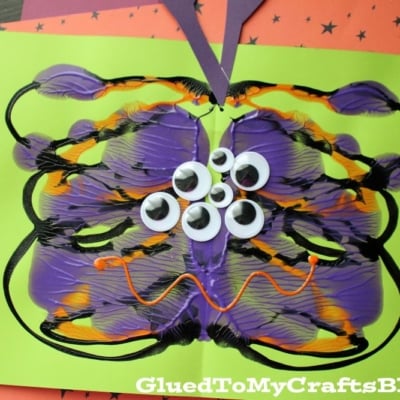 A colorful craft features a symmetrical puffy paint splat monster with purple, orange, and black paint, several googly eyes, and purple paper antennae on green paper, set on a star-patterned background.