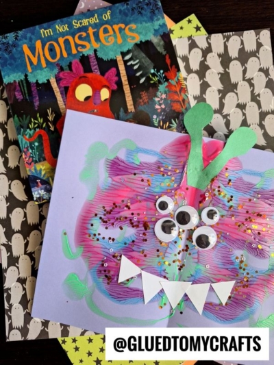 A colorful homemade puffy paint splat monster craft with googly eyes and white paper teeth sits next to an “I’m Not Scared of Monsters” book on ghost-patterned paper. @gluedtomycrafts is written at the bottom.