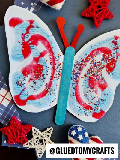 Patriotic paint splatter butterfly craft made from white paper with blue and red paint, star-shaped glitter, and craft sticks. The butterfly is surrounded by star decorations and plaid paper on a dark background.