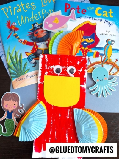 Colorful pirate and ocean-themed children’s craft scene with books, a paper mermaid, octopus, bird made from cupcake liners, a pink fish cutout, and a fun paper bag parrot kid craft. Books include “Pirates Love Underpants” and “Pete the Cat.”.