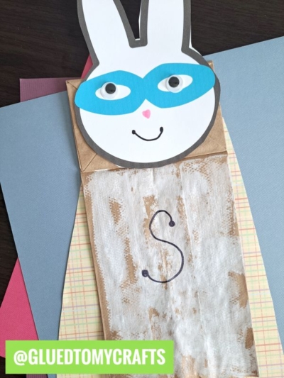 A bunny superhero paper bag puppet with a blue mask, googly eyes, and a drawn “S” on its chest sits on colorful paper sheets. The handle @GLUEDTOMYCRAFTS is shown at the bottom. Perfect for your own lunchsack superhero bunny craft!.