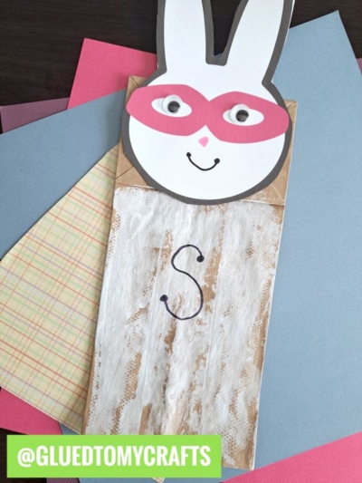 A handmade paper bag superhero bunny puppet with a pink mask, googly eyes, and a drawn smile features the letter S on its body. It’s placed on colored paper sheets. @GLUEDTOMYCRAFTS is written at the bottom.