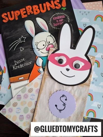 A children’s book titled SUPERBUNS! with a bunny on the cover lies beside a handmade paper bag Easter bunny superhero puppet, sporting a pink mask, googly eyes, and a dollar sign on its chest, with colorful paper in the background.
